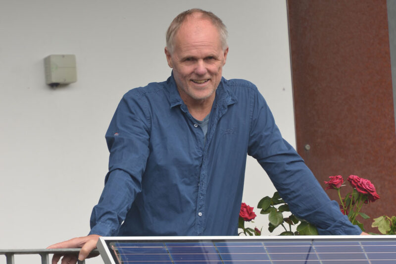 Jörg Lange, ein Mann im "besten Alter" stützt sich auf ein Balkongeländer, an dem ein Photovoltaikmodul als Balkonkraftwerk befestigt ist.