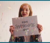 Im Bild eine Frau mit einem Schild „eine spektakuläre Idee“, die für Klima° vor acht wirbt.