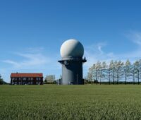 Flaches Feld mit Haus und einem niedrigen Turm mit einer weißen Kugel. Radar-Anlage als Symbol für Flächenkonflikt von Windenergie und Militär.