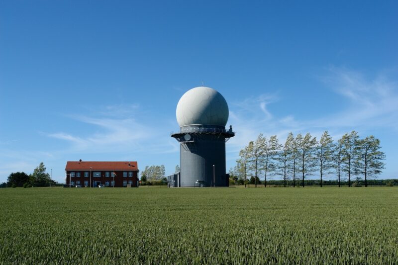 Flaches Feld mit Haus und einem niedrigen Turm mit einer weißen Kugel. Radar-Anlage als Symbol für Flächenkonflikt von Windenergie und Militär.