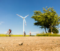 Drei Menschen auf dem Fahrrad fahren vor einer Windenergie-Anlage vorbei - Symbolbild für die Energiewende-Radtour