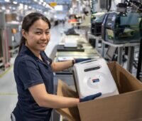 Eine Frau in einer Fabrik präsentiert eine Box mit einem Ladegerät.