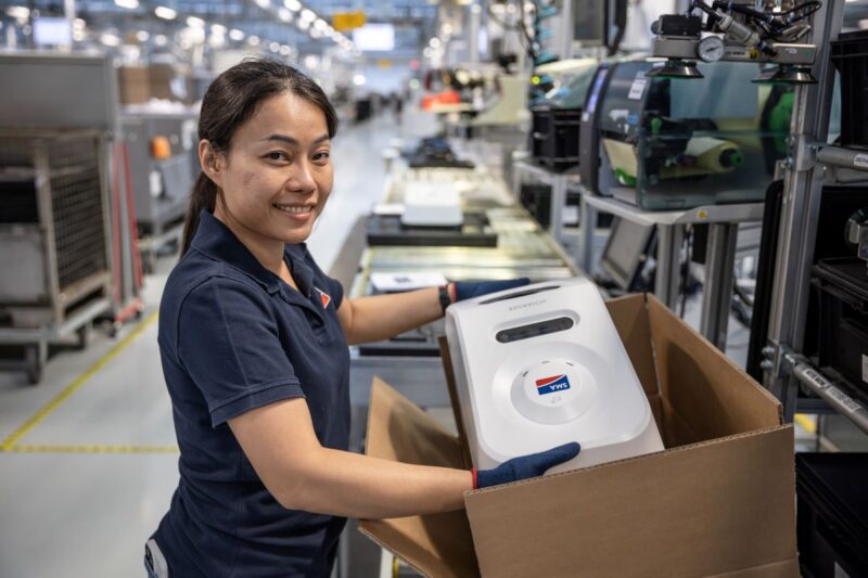 Eine Frau in einer Fabrik präsentiert eine Box mit einem Ladegerät.