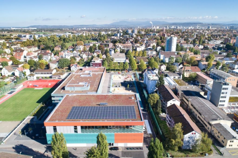 Luftaufnahme des Schweizer Ortes Allschwil. Im Vordergrund eine Schulturnhalle mit einer PV-Anlage auf dem Flachdach.