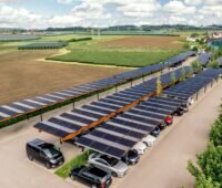 Luftaufnahme von parkenden Autos unter Solar-Carports am Ortsrand neben Feldern.