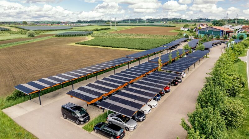 Luftaufnahme von parkenden Autos unter Solar-Carports am Ortsrand neben Feldern.