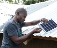 Ein Afrikaner montiert ein Solarmodul vor zwei Hütten mit Wellblechdach.
