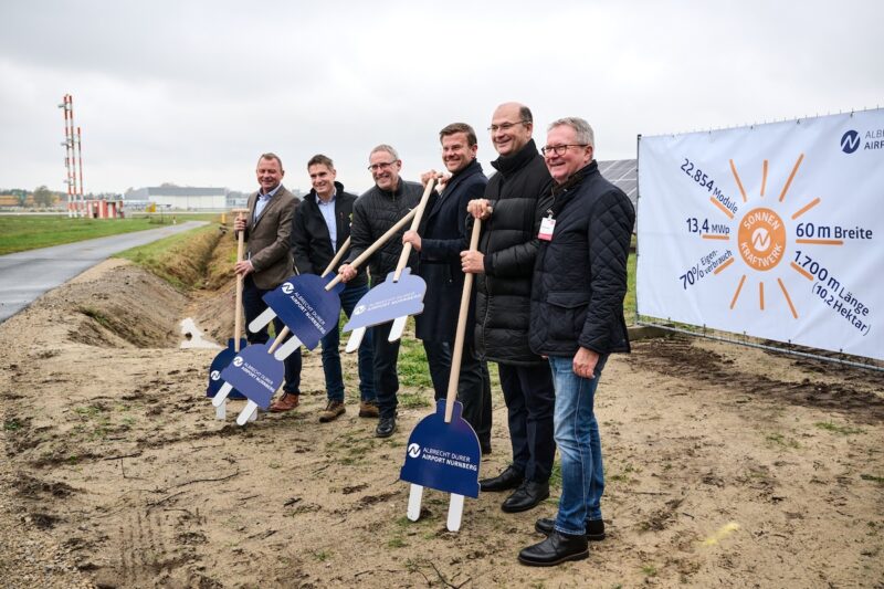 Menschen mit Grabwerkzeugen die aussehen wie Schuko-Stecker an Stöcken - Spatenstich am Flughafen Nürnberg für Solaranlage.