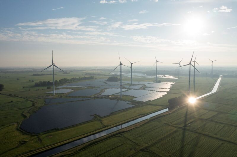 Luftbild zeigt großen PV-Park unter Windenergieanlagen im grünen Flachland.