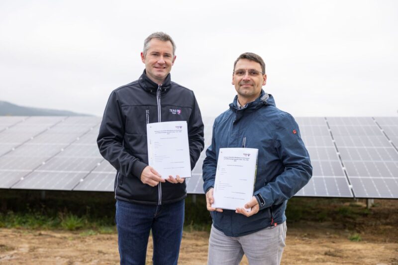 Zwei Männer mit je einem Vertragsexemplar in den Händen vor einer Freiflächensolaranlage.