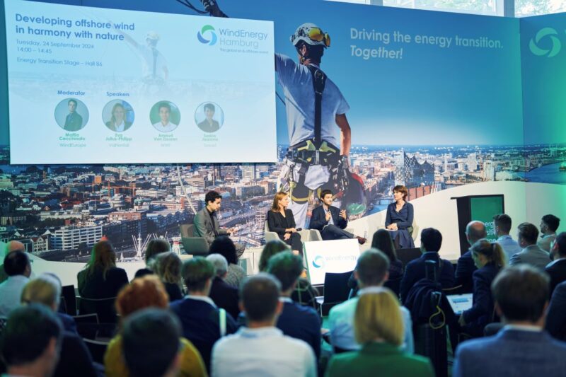 Konferenz mit vier Personen auf dem Podium und Fotos der Windenergie.