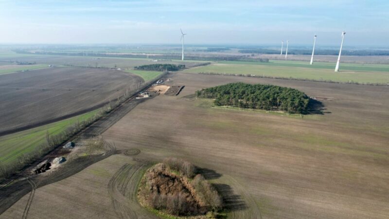 Blick über flaches Land auf einige Windenergie-Anlagen mit hohem Turm und kleinem Rotor.
