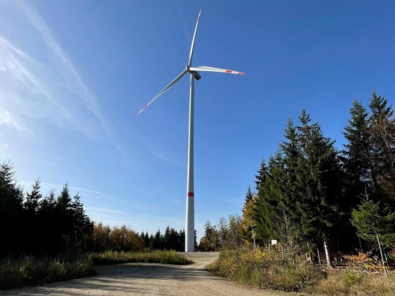 Windenergie-Anlage vor blauem Himmel, davor ein Weg und Wald aus Nadelbäumen.