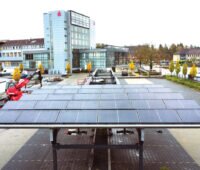 Solarcarport auf einem Parkplatz. Im Hintergrund das Gebäude der Sparkasse.