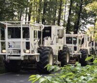 Weiß lackierte Kraftwagen mit großen Reifen in einer Reihe auf einem Waldweg. Vibro-Trucks erkunden den Untergrund, um das Potenzial für Tiefengeothermie-Bohrungen zu erkunden.