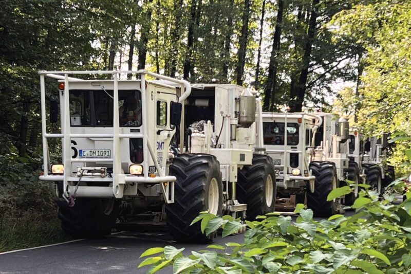 Weiß lackierte Kraftwagen mit großen Reifen in einer Reihe auf einem Waldweg. Vibro-Trucks erkunden den Untergrund, um das Potenzial für Tiefengeothermie-Bohrungen zu erkunden.