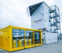 Gelber Container mit Glasfront, dahinter ein grauer Turm - Anlage für Solar-Kraftstoff-Herstellung von Synhelion.